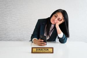 junge kaukasische brünette geschäftsfrau direktorin im büro am schreibtisch sitzen telefon scrollen nachrichten social media gelangweilt bei der arbeit direktor lustig. unnötige Mitarbeiterbüro-Streichplatte foto