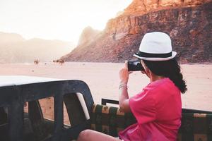 Touristin bei Sonnenuntergang Machen Sie ein Smartphone-Foto von Kamelen mit Reitern von einem fahrenden 4WD-LKW auf einer Wüsten-Sonnenuntergangstour in der Wadi-Rum-Wüste im Freien. Passagiersicht foto