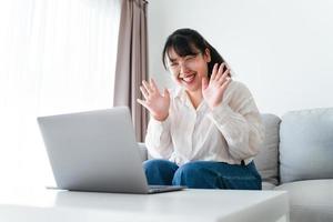 junge asiatin, die einen laptop für online-videokonferenzen verwendet, winkt mit der hand und macht eine hallo-geste auf der couch im wohnzimmer. foto