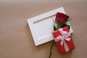 Blick von oben auf die rote Rose und die rote Geschenkbox, die auf einem weißen, leeren Fotorahmen liegen, der auf braunem Hintergrund isoliert ist. valentinstag, hochzeit, geburtstag und konzept für besondere anlässe. flache Lage, Draufsicht. foto