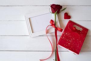 Rote Rose mit rotem Band auf einem weißen leeren Fotorahmen und roter Geschenktüte auf weißem Holzhintergrund. konzept für valentinstag, geburtstag und besondere anlässe. flache Lage, Draufsicht. foto