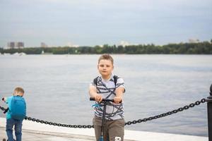 ein Junge auf einem Roller am Ufer der Stadt entlang. Reise. Rucksack auf dem Rücken. Das Gesicht drückt natürliche freudige Emotionen aus. nicht inszenierte Fotos aus dem Leben