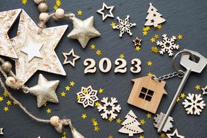 Hausschlüssel mit Schlüsselanhänger Häuschen auf schwarzem Hintergrund mit Sternen, Schneeflocken. frohes neues jahr 2023-holzbuchstaben, grußkarte. Kauf, Bau, Umzug, Hypothek foto