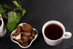 Weiße Keramiktasse mit heilendem Aufguss aus Birkenpilz-Chaga, Schüssel mit Chaga-Stücken und Birkenblättern auf Schwarz. foto