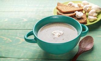 Türkisfarbene Keramikschale mit Pilzcremesuppe, Holzlöffel, Knoblauch, Brot, auf grünem Holzhintergrund. foto