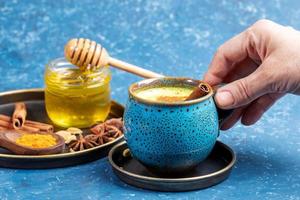 weibliche hand, die tasse traditionelles ayurvedisches getränk goldene kurkumamilch und teller mit seinen zutaten auf blau hält. foto