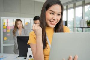 junge asiatische glückliche geschäftsfrau oder praktikantin, die computer im modernen bürohintergrund verwendet, ist kollegen, die mit einer neuen startprojektidee diskutieren. Befähigter digitaler Unternehmer arbeitet an Startup-Projekt