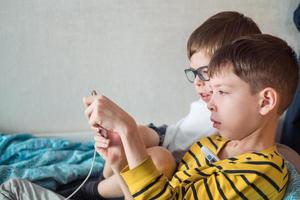 Zwei Jungen liegen krankgeschrieben mit einem Thermometer unter der Achselhöhle und einem Telefon in der Hand auf dem Bett. kranke kinder zu hause foto