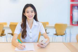 junge asiatische studentin in uniform, die smartphone benutzt und etwas über die arbeit schreibt. es gibt viele dokumente auf dem tisch, ihr gesicht mit einem lächeln in einem arbeiten, um informationen für einen studienbericht zu suchen