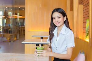 porträt einer süßen asiatischen thailändischen studentin in uniform sitzt glücklich lächelnd und selbstbewusst erfolgreich, während sie ein glas getränk in einem café in der universität hält. foto