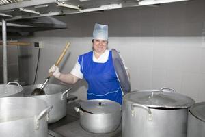 Eine Frau kocht in einer Industrieküche mit einer großen Kelle zwischen Metallpfannen. Koch im Krankenhaus foto