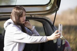 Ein schönes Mädchen sitzt in einem Auto und schaut auf den Telefonbildschirm. ein Selfie in der Natur machen oder streamen. foto