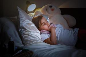 Das kranke kleine Mädchen, das mit einer Decke bedeckt ist, umarmt den Teddybären und schaut traurig auf die Medizin, während es liegt foto