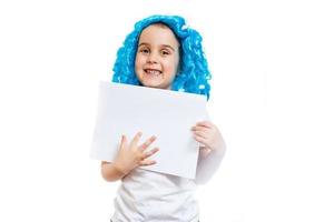 junges kleines Mädchen in blauer Perücke, das weißes leeres Blatt Papier lokalisiert auf Weiß hält foto