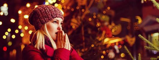 Outdoor-Foto von jungen schönen glücklichen lächelnden Mädchen, die auf der Straße posieren. festliche weihnachtsmesse im hintergrund. Modell mit stylischem Wintermantel, Strickmütze, Schal. foto