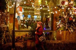 Outdoor-Foto von jungen schönen glücklichen lächelnden Mädchen, die auf der Straße posieren. festliche weihnachtsmesse im hintergrund. Modell mit stylischem Wintermantel, Strickmütze, Schal. foto