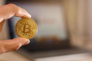 Bitcoin-Goldmünze in der Hand des Menschen auf dem Hintergrund des Laptops auf einem weißen Tisch