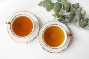 Auf dem Tisch stehen zwei Teetassen, ein Eukalyptuszweig. schwarzer Tee in einer schönen weißen Porzellantasse mit goldenem Schliff.