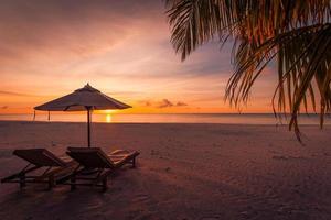 perfekter strand sonnenuntergang, tropischer sonnenuntergang mit zwei stühlen romantische aussicht mit palmblättern. schöne Strandnatur. luxusresort oder hotelhintergrund. Blick auf den Sonnenuntergang, Sonnenstrahlen und entspannende Stimmung, Urlaubsstimmung foto