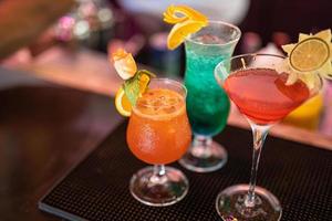 exotische cocktails, mojito-cocktail, orangencocktail, erdbeercocktail in glasgläsern mit strohhalmen und früchten und verschwommenem barzubehör shaker, löffel, würze bunte cocktails auf der bartheke foto