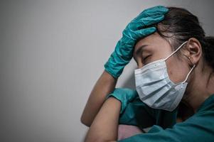 müde depressive asiatische krankenschwester trägt gesichtsmaske blaue uniform sitzt auf dem krankenhausboden, junge ärztin gestresst von harter arbeit foto