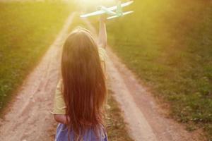 glückliches Kindermädchen mit langen blonden Haaren, das bei Sonnenuntergang mit Spielzeugflugzeug im Freien spielt foto