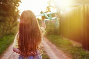 glückliches Kindermädchen mit langen blonden Haaren, das bei Sonnenuntergang mit Spielzeugflugzeug im Freien spielt foto