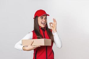 Bild einer aufgeregten, glücklichen jungen Lieferfrau in roter Kappe, die mit einem Paketbriefkasten steht, der über weißem Hintergrund isoliert ist. suchende Kamera. foto