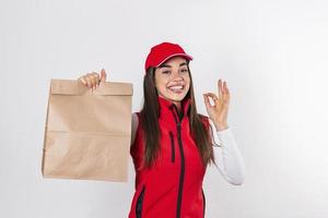 lieferfrau in roter uniform hält kraftpapierpaket mit lebensmitteln isoliert auf weißem hintergrund, studioportrait. weibliche angestellte im kappen-t-shirt-druck, die als kurier arbeitet. Service-Konzept. foto