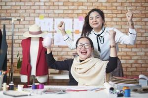 asiatische modedesignerin mittleren alters und junge jugendliche auszubildende schneiderin feiern und fröhlich mit dem erfolg der kleiderdesignarbeit und befriedigen im stylistenstudio, glückliches kostümboutique-kleinunternehmen. foto