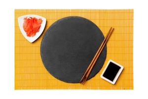 leere runde schwarze Schieferplatte mit Essstäbchen für Sushi, Ingwer und Sojasauce auf gelbem Bambusmattenhintergrund. draufsicht mit kopierraum für ihr design foto