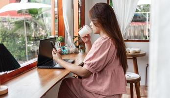 eine glückliche frau in einem café mit einem smartphone und einem laptop, der kaffee in einem pappbecher im café trinkt. Junge weiße Frau mit langen Haaren, die in einem Café sitzt und an ihrem Smartphone arbeitet.