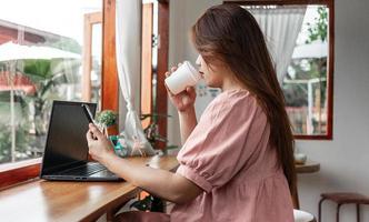 eine glückliche frau in einem café mit einem smartphone und einem laptop, der kaffee in einem pappbecher im café trinkt. Junge weiße Frau mit langen Haaren, die in einem Café sitzt und an ihrem Smartphone arbeitet.