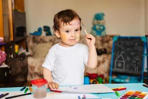 Ein kleiner süßer Junge zeichnet mit Pinseln und farbigen Farben auf ein Blatt Papier foto