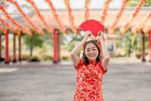 glückliche junge dame, die traditionelles cheongsam qipao kostüm trägt, das ang pao, rote umschläge im chinesischen buddhistischen tempel hält. Feiern Sie das chinesische Neujahrsfest, die Feiertage der festlichen Jahreszeit foto