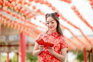 schöne asiatische frau, die glücklich lächelt und ang pao hält, rote umschläge, die cheongsam tragen und selbstbewusst im chinesischen buddhistischen tempel aussehen. Feiern Sie das chinesische Neujahrsfest, die Feiertage der festlichen Jahreszeit. foto