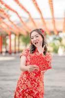 glückliche schöne asiatische frau, die traditionelles cheongsam qipao kostüm trägt, das ang pao, rote umschläge im chinesischen buddhistischen tempel hält. chinesischer text bedeutet viel glück, großer gewinn foto