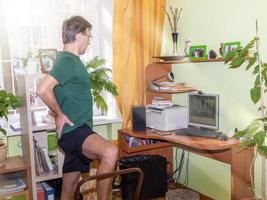 Reifer Mann probt Yoga-Asanas mit Stuhl, der sorgfältig auf den Computermonitor im sonnigen Zimmer zu Hause schaut. Fernsport. foto