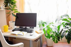 echter heimarbeitsplatz mit tisch, ausgeschaltetem computer, grünen zimmerpflanzen, gelber tasse und notizblock in der nähe des fensters. foto