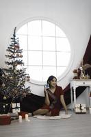 porträt des hübschen jungen mädchens, das sich gemütlich hinsetzt und weihnachtsgeschenk hält, lächelndes rotes kleid im geschmückten weihnachtswohnzimmer drinnen trägt foto