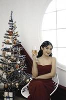 porträt eines hübschen jungen mädchens, das sich gemütlich hinsetzt und wein trinkt, trägt ein rotes kleid in einem dekorierten weihnachtswohnzimmer drinnen foto