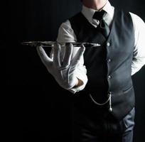Porträt eines Kellners oder Butlers in weißen Handschuhen, der ein silbernes Serviertablett auf schwarzem Hintergrund hält. konzept der dienstleistungsbranche und der professionellen höflichkeit. foto