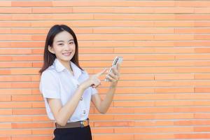 Porträt einer süßen asiatischen thailändischen Studentin in Uniform steht glücklich lächelnd und selbstbewusst erfolgreich, während sie ein Smartphone mit Ziegelwänden als Hintergrund in der Universität hält. foto