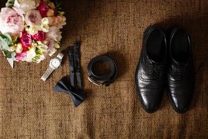 Schwarze Schuhe, schwarzer Gürtel, schwarze Uhr, schwarzer Schmetterling, Manschettenknöpfe und Parfüm auf braunem Hintergrund mit Sackleinen foto