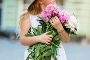 Kleines Mädchen, das Pfingstrosenblumenstrauß riecht, sitzt in einem weißen Stuhl im sonnigen Schlafzimmer. Kinderzimmereinrichtung mit Blumen für Mädchen. Kind bereit für Geburtstagsfeier mit rosa Pfingstrosen. kinder spielzimmer. foto