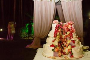 Auf dem weißen Tisch in der Mitte des Restaurantsaals steht eine originelle Hochzeitstorte inmitten von Blumen foto