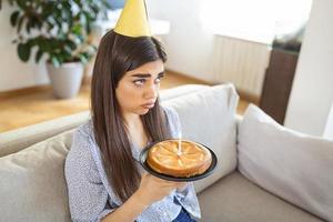 virtuelle Party während des Lockdowns. verärgerte und frustrierte alleinstehende afrikanisch-amerikanische Frau mit Partyhut, die online Geburtstag feiert. dame, die videokonferenz macht, kuchen mit kerze hält, auf dem boden sitzt foto