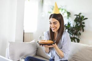 virtuelle Party. glückliche junge frau mit hut, die sich per videokonferenz online mit freunden und familie trifft, einen kuchen hält, anstößt und geburtstag feiert, zu hause bleibt foto