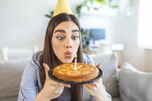 neues normales Konzept. glückliche frau, die ihren geburtstag allein feiert. junge Frau, die einen Kuchen hält. Geburtstagsfeier zu Hause. Videoanruf für die Familie. soziale Distanzierung. Leben zu hause. einen Wunsch machen foto