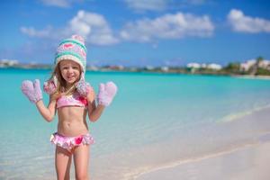 Kleines entzückendes Mädchen in warmer Strickmütze und Fäustlingen an einem tropischen Strand foto